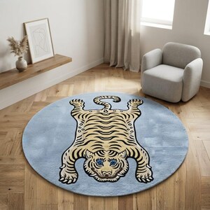 Può includere: Un tappeto circolare con uno sfondo azzurro e una grafica centrale di una tigre stilizzata. La tigre è raffigurata in toni crema e marrone chiaro con strisce marrone scuro, una coda arricciata e occhi blu brillante al centro del tappeto.