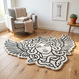 Op de afbeelding: Een crèmekleurig vloerkleed met een zwarte lijntekening van een gezicht met slangen als haar en grote vleugels die aan de zijkanten uitsteken. Het kleed heeft een aangepaste vorm die de contouren van het gevleugelde ontwerp volgt.