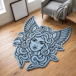 Op de afbeelding: Een lichtblauw vloerkleed in de vorm van het hoofd van Medusa met zwarte lijntekeningen. Het ontwerp toont een centraal gezicht omringd door een bos slangen als haar en grote vleugels aan de zijkanten, wat een opvallend silhouet creëert.