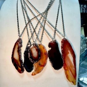 May include: A display of necklaces featuring polished agate pendants in shades of brown, orange, and black. Each pendant is unique in shape and pattern, suspended from a silver chain. The necklaces are displayed on a white mannequin.