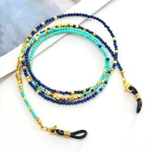 May include: Eyeglass chain featuring three strands of beads in turquoise, navy blue, and gold. The chain has a gold clasp and black rubber loops for attaching to glasses. The chain also has a decorative black horn charm.
