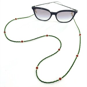 May include: Black cat-eye sunglasses with a green beaded eyeglass chain. The chain features small green beads and red accent beads. The glasses have a gradient lens.