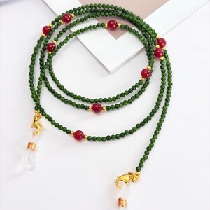 May include: A green beaded eyeglass chain with red accent beads and gold-colored clasps. The chain is made of small, round green beads and features larger red beads spaced along its length. The clasps are designed to hold eyeglasses.