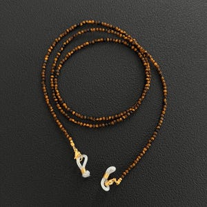 May include: A beaded eyeglass chain with small, round, brown and black beads. The chain has gold-colored metal accents and white rubber loops to attach to glasses. The chain is displayed on a black surface.