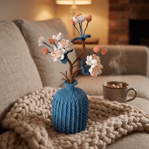 Op de afbeelding: Een blauwe gebreide vaas bevat een gehaakte bloemstuk met witte en perzikbloesems. De vaas staat op een dikke gebreide deken. Een mok met warme drank staat op de achtergrond.