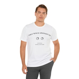 Puede incluir: Camiseta blanca con el texto "THEY WALK AMONGST US" sobre un gráfico de cara simple con dos ojos y una línea para la boca. Debajo de la cara, el texto dice "and they send emails". La camiseta la lleva una persona.