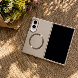 May include: A champagne-colored foldable smartphone case featuring a circular magnetic charging ring on the back panel, a dual-lens camera module with a white housing, and a black screen display visible on the front section of the device.