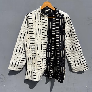 May include: A long-sleeved quilted jacket featuring a high collar and a button-down front. The garment displays a contrasting black and white pattern of vertical lines and dots. It includes a single patch pocket on the lower front section.