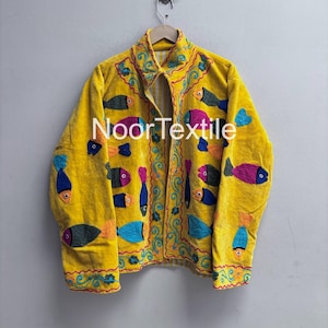 May include: A yellow velvet jacket with colorful embroidered fish. The jacket has a collared neckline and is decorated with blue, pink, orange, and black fish. The text "NoorTextil" is visible on the front.