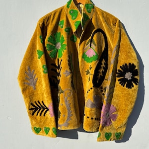 Könnte beinhalten: Senfgelbe Samtjacke mit gestickten Blumen- und Paisley-Designs in Grün, Schwarz, Rosa und Grau. Die Jacke hat einen Reißverschluss vorne und einen Kragen mit grünen Stickereidetails.