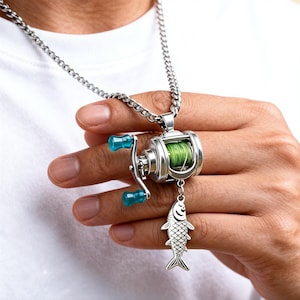 Puede incluir: Collar con colgante de carrete de pesca plateado con cadena. El carrete tiene asas turquesas y línea verde. Un dije de pez plateado cuelga debajo. El collar se usa sobre una camiseta blanca.