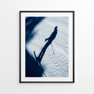 Peut inclure: Impression encadrée présentant l'ombre bleu foncé d'un skieur sur une pente enneigée. Le skieur tient des bâtons de ski, et l'image a une qualité artistique à fort contraste. L'impression est dans un cadre noir.