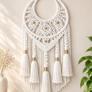 May include: A white macrame wall hanging featuring a circular frame with intricate knotted patterns, three gold-toned circular accents, and five long hanging tassels with gold-toned cylindrical beads at the top of each tassel, suspended by a cord.