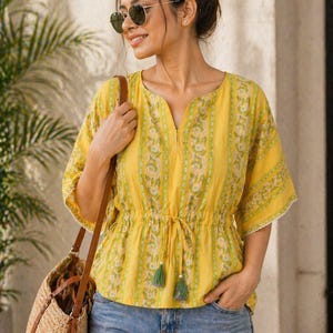 Puede incluir: Una blusa amarilla con estampado floral y cintura con cord&oacute;n. La blusa tiene mangas cortas con ribete de encaje y se combina con vaqueros azules. Se lleva un bolso de paja tejido con correas de cuero marr&oacute;n al hombro.