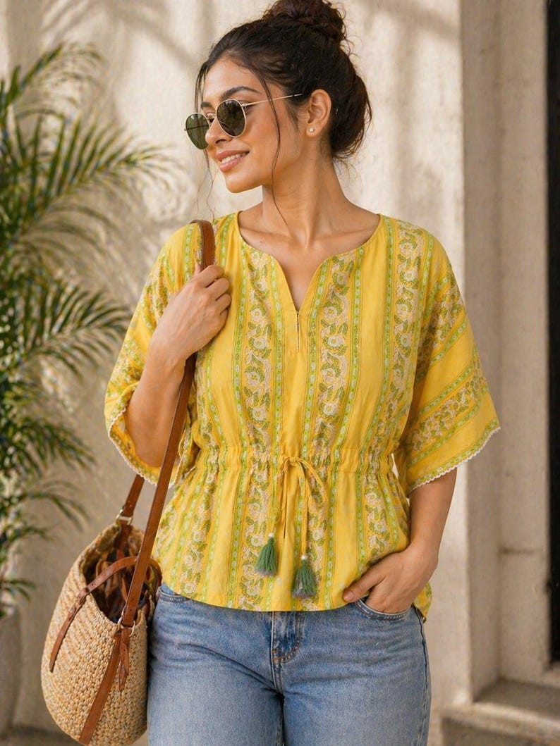 Puede incluir: Una blusa amarilla con estampado floral y cintura con cord&oacute;n. La blusa tiene mangas cortas con ribete de encaje y se combina con vaqueros azules. Se lleva un bolso de paja tejido con correas de cuero marr&oacute;n al hombro.