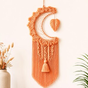 May include: A crescent moon-shaped macrame wall hanging in a vibrant peach colour. The piece features intricate knotted patterns, a central leaf-shaped element suspended within the curve, and a long fringe base with a central tassel detail.