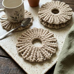 May include: A set of round, beige macrame coasters featuring a knotted, radial pattern with a central circular opening. The coasters are crafted from thick cotton cord and are arranged on a speckled, off-white ceramic tray alongside a mug.