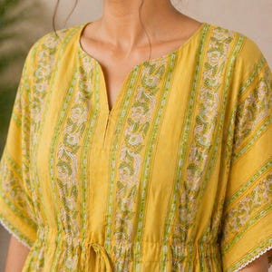 Puede incluir: Vestido t&uacute;nica amarillo con cuello en V y cintura con cord&oacute;n. El vestido presenta rayas verticales con un estampado floral en verde y blanco. Las mangas tienen ribete de encaje blanco y el cord&oacute;n tiene borlas verdes.
