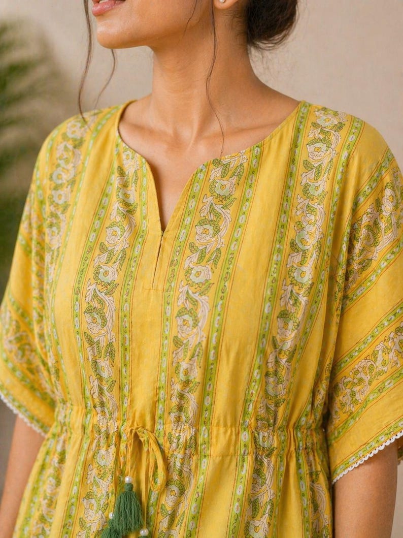 Puede incluir: Vestido t&uacute;nica amarillo con cuello en V y cintura con cord&oacute;n. El vestido presenta rayas verticales con un estampado floral en verde y blanco. Las mangas tienen ribete de encaje blanco y el cord&oacute;n tiene borlas verdes.