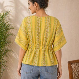 Puede incluir: Una blusa amarilla con estampado floral y cintura ce&ntilde;ida. La blusa tiene mangas cortas con ribete de encaje blanco y cuello en V. La modelo lleva vaqueros azules. El fondo incluye una planta y una silla.