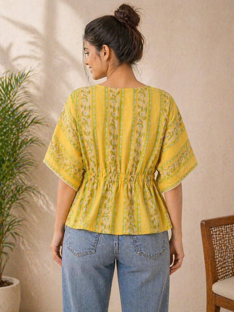 Puede incluir: Una blusa amarilla con estampado floral y cintura ce&ntilde;ida. La blusa tiene mangas cortas con ribete de encaje blanco y cuello en V. La modelo lleva vaqueros azules. El fondo incluye una planta y una silla.