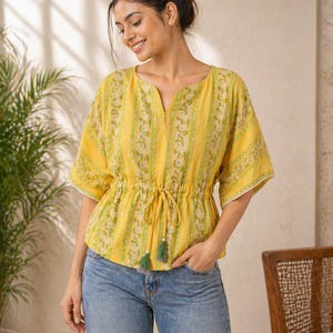 Puede incluir: Top amarillo con estampado floral y cintura con cord&oacute;n. La parte superior tiene mangas cortas con ribete de encaje blanco y cuello en V. La modelo lleva vaqueros azules. El top tiene borlas verdes.