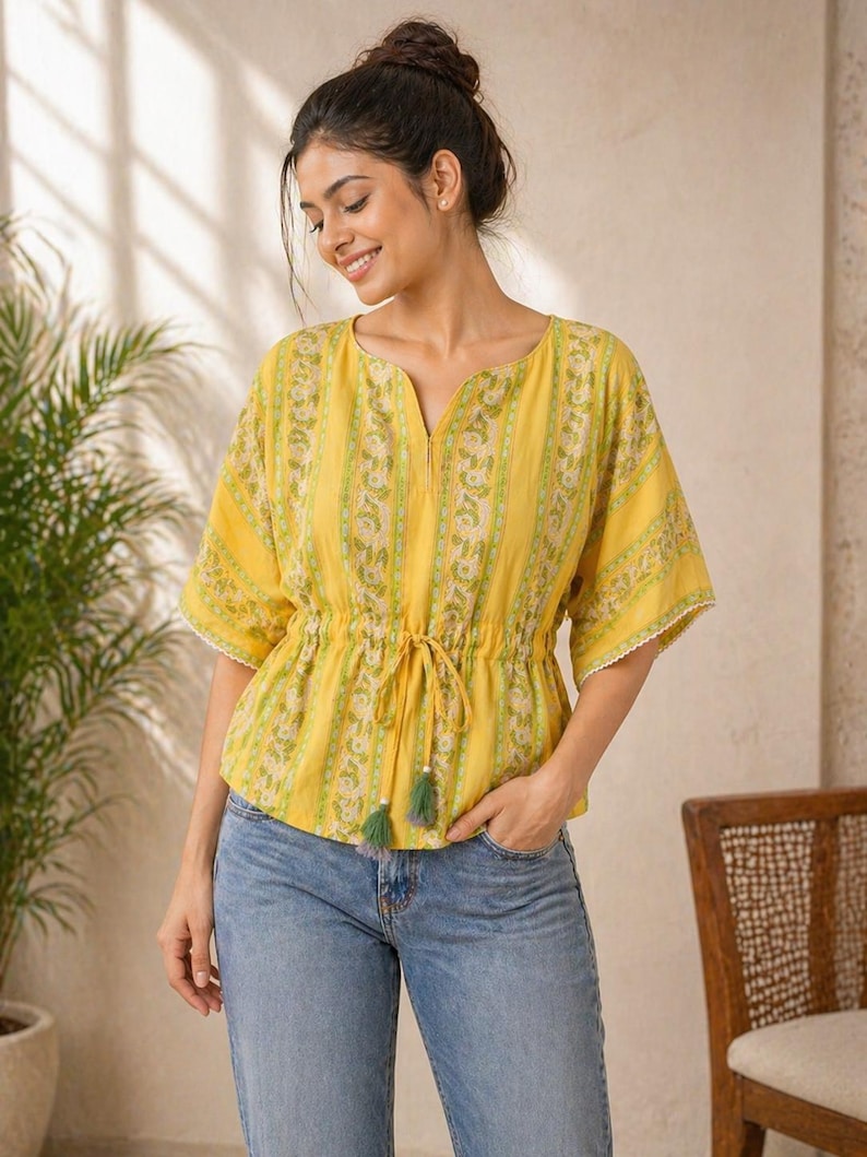 Puede incluir: Top amarillo con estampado floral y cintura con cord&oacute;n. La parte superior tiene mangas cortas con ribete de encaje blanco y cuello en V. La modelo lleva vaqueros azules. El top tiene borlas verdes.