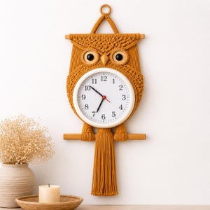 May include: A mustard-coloured macrame owl clock. The clock has a white face with black numerals and hands, and a red second hand. The owl has large, round black eyes and a tassel tail. The clock is designed to be hung on a wall.