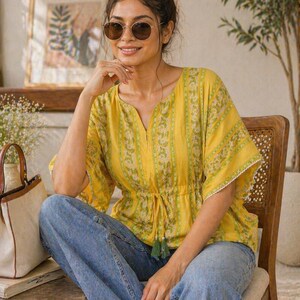 Puede incluir: Una mujer con un top amarillo con estampado floral y vaqueros azules. El top tiene cintura con cord&oacute;n y mangas cortas con ribete de encaje blanco. Lleva gafas de sol y est&aacute; sentada en una silla de madera.