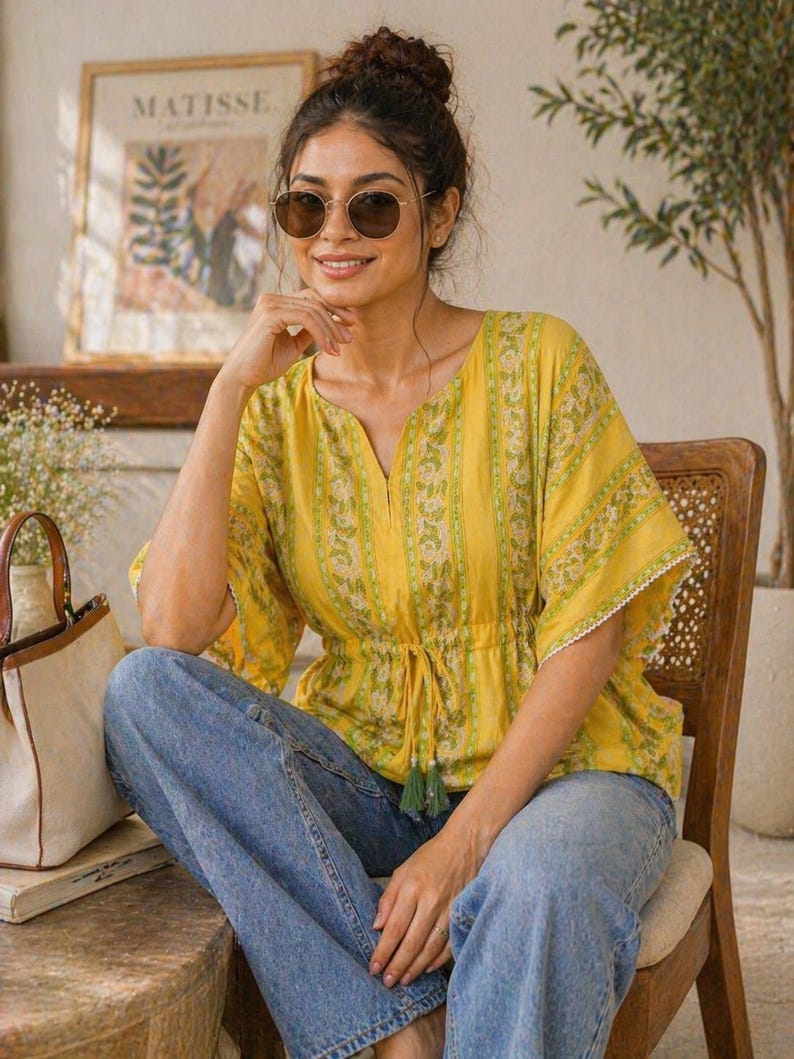 Puede incluir: Una mujer con un top amarillo con estampado floral y vaqueros azules. El top tiene cintura con cord&oacute;n y mangas cortas con ribete de encaje blanco. Lleva gafas de sol y est&aacute; sentada en una silla de madera.
