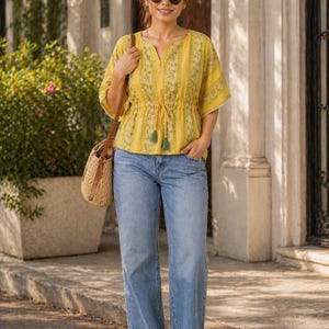 Puede incluir: Una mujer con un top amarillo con estampado floral y vaqueros azules. El top tiene mangas cortas y cintura ajustable. Lleva gafas de sol y un bolso de paja. El conjunto es informal y adecuado para el clima c&aacute;lido.