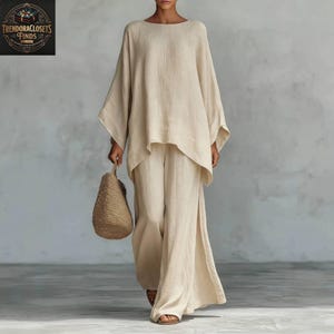 Op de afbeelding: Een beige linnen tweedelige outfit. De top is een loszittende tuniek met lange mouwen en een boothals. De broek heeft wijde pijpen met een split aan de zijkant. Een geweven beige tote bag wordt in de hand gehouden. De outfit is casual en comfortabel.