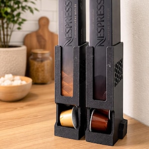 May include: Black coffee pod holder with two compartments, each holding a Nespresso box. The boxes are black with the Nespresso logo and product names. The holder is on a wooden surface, with a blurred background.