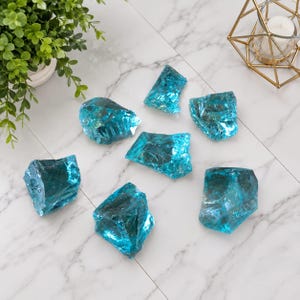 May include: Seven irregular, translucent turquoise blue glass chunks with jagged, crystalline edges and a reflective, glossy surface. These decorative glass pieces are scattered across a white surface, showcasing varying shapes and sizes with internal fractures.