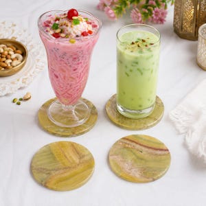 May include: Two round green stone coasters sit on a white surface. One coaster supports a tall glass filled with a pink layered dessert topped with a cherry, while the other holds a tall glass containing a light green beverage with green bits.