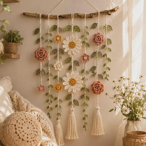 May include: A wall hanging featuring crocheted flowers and leaves suspended from a natural wooden branch. The piece includes white daisies, pink and yellow roses, and green vines, with three cream-colored yarn tassels hanging from the bottom.