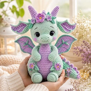 May include: A crocheted dragon plush toy featuring a light green body with a purple belly, wings, and tail spikes. The dragon has large black eyes, pink cheeks, and is decorated with small purple and light green crocheted flowers on its head and body.