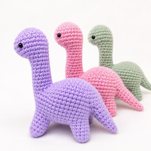 May include: Three crocheted dinosaur toys. The first is purple, the second is pink, and the third is green. Each has a long neck, small black eyes, and four legs. The toys are handmade and suitable for children.