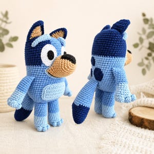 May include: Two crochet figures of a blue dog with black and tan accents. The dog has large black eyes, a tan nose, and a light blue belly. The figure is standing upright with its arms and legs extended. The second figure is a side view of the same dog.