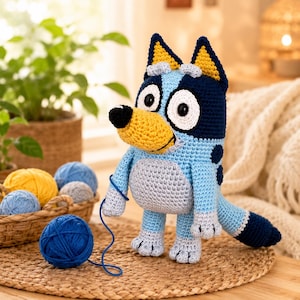 May include: A handmade crochet dog toy in blue, yellow, and gray. The dog has large white eyes, a yellow snout, and a black nose. A blue ball of yarn is in front of the dog, and a basket of yarn balls is in the background.
