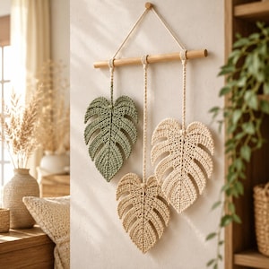 May include: A wall hanging featuring three crocheted monstera leaves in olive green, beige, and cream. The leaves are suspended by braided cords from a horizontal wooden dowel, which is attached to a single piece of string for wall mounting.