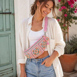 May include: A pink and multi-colored crochet fanny pack with a matching strap. The bag features a floral design with shades of pink, green, and orange. A small, round, matching charm with a tassel hangs from the strap. The bag is worn across the body.