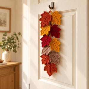 May include: A vertical wall hanging decoration featuring a series of crocheted maple leaves in autumn colors including deep red, bright yellow, burnt orange, and muted brown, all connected by a central string and suspended from a black door hook.