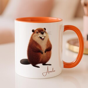 Può includere: Tazza in ceramica bianca con interno e manico arancione. La tazza presenta un'illustrazione di un castoro dei cartoni animati con pelliccia marrone e un'espressione sorridente. Il nome "Amelie" è scritto in corsivo sulla tazza.