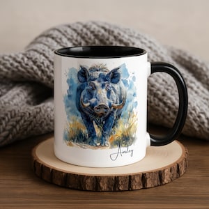 Pode incluir: Caneca de cerâmica branca com alça e interior pretos. A caneca apresenta uma ilustração em aquarela de um javali azul com grandes presas, em um fundo azul e amarelo. O nome "Ainsley" está escrito em itálico na caneca.