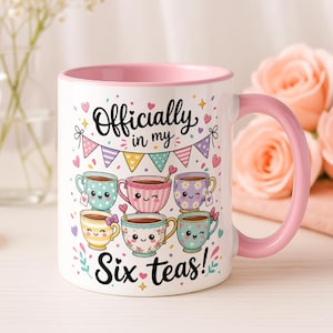 May include: White ceramic mug with a pink interior and handle. The mug features the text "Officially in my Six teas!" and illustrations of six smiling teacups with different colours and designs. The mug is decorated with hearts and confetti.