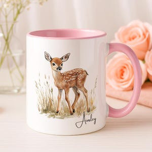 May include: White ceramic mug with a pink interior and handle. The mug features a watercolour illustration of a fawn with brown spots and the name "Ainsley" in cursive. The fawn is standing in tall grass.