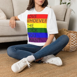 Può includere: Una t-shirt bianca a maniche corte con una grafica rettangolare a strisce orizzontali rosse, arancioni, gialle, verdi, blu e viola. Il testo "LOVE WHOEVER THE FUCK YOU WANT" è stampato in lettere maiuscole nere sopra le strisce.