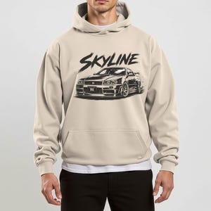Könnte beinhalten: Beige Kapuzenpullover mit einer schwarzen Grafik eines Skyline-Autos und dem Wort "Skyline" darüber. Der Hoodie hat eine Kapuze mit Kordelzug und eine Kängurutasche.