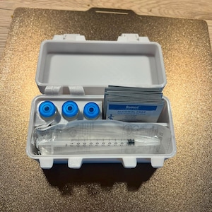 Op de afbeelding: Een witte plastic opbergdoos met drie kleine flesjes met blauwe doppen, een stapel Romed alcoholdoekjes en een enkele doorzichtige plastic injectiespuit in een verzegelde verpakking. De doos staat open en toont de georganiseerde medische benodigdheden.